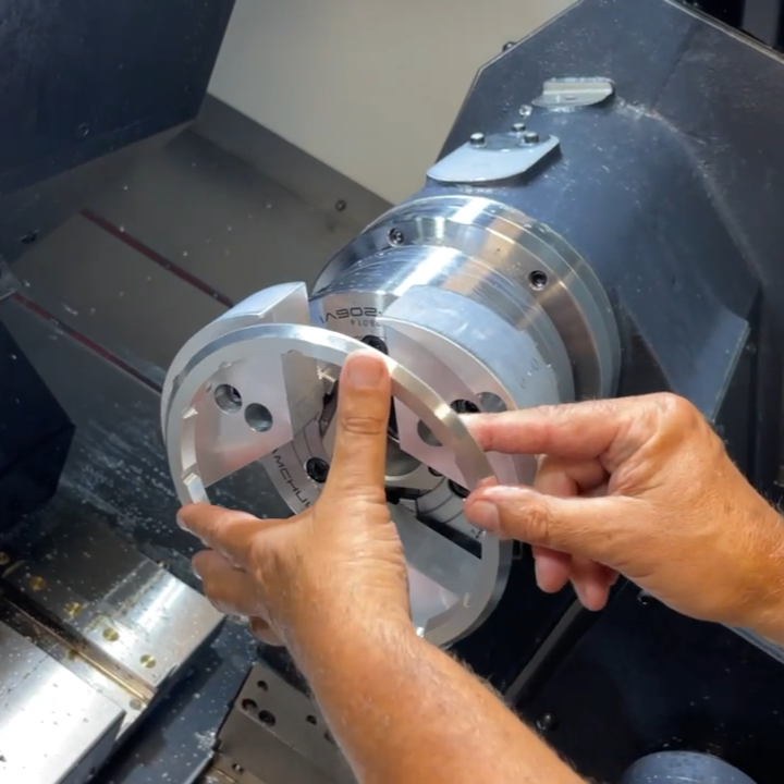 In-House Aluminum CNC Machining Hands positioning a machined aluminum ring on a CNC lathe chuck inside a machining enclosure.
